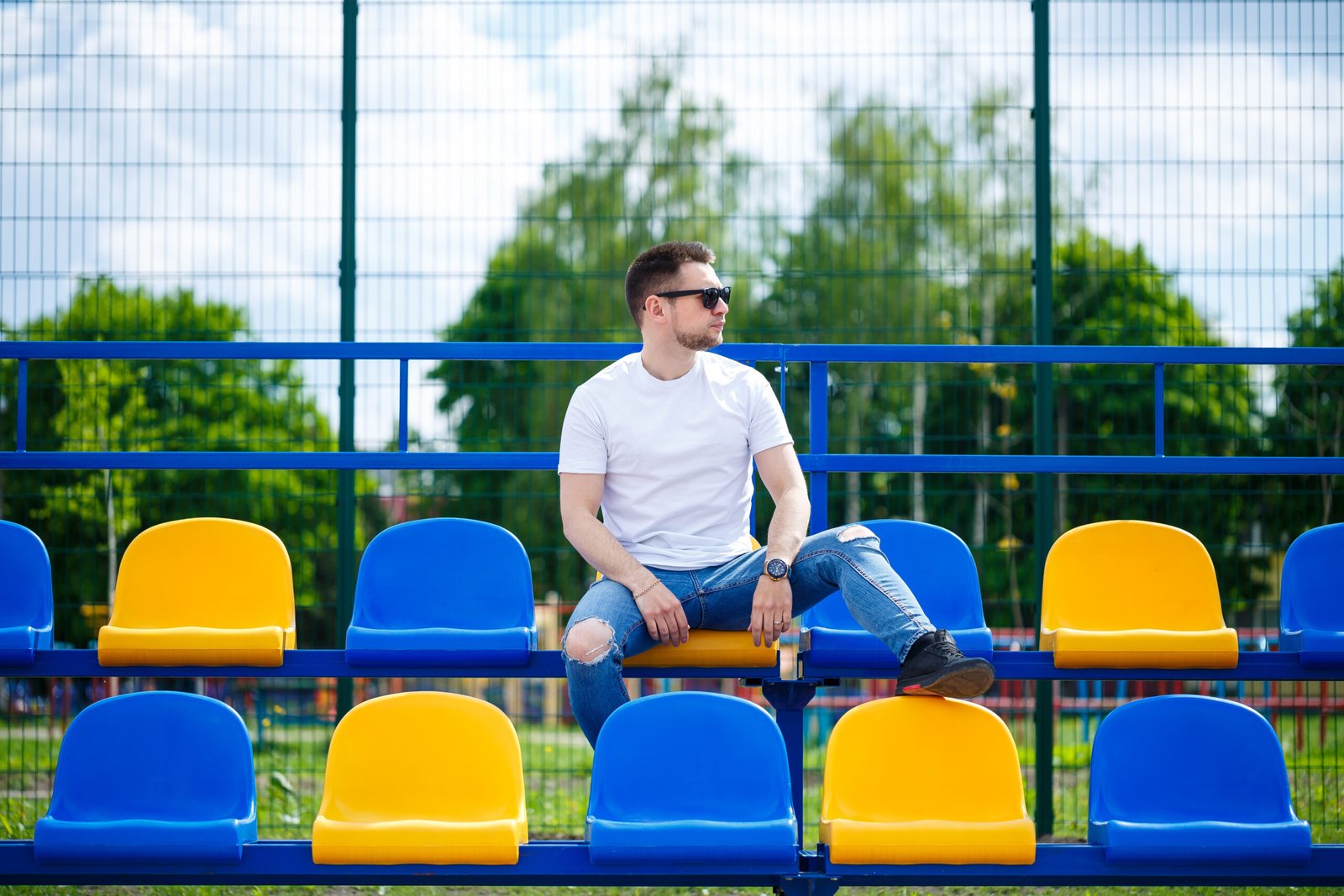 A guy in a white T-shirt sits in a stadium on the chairs for the fans alone.