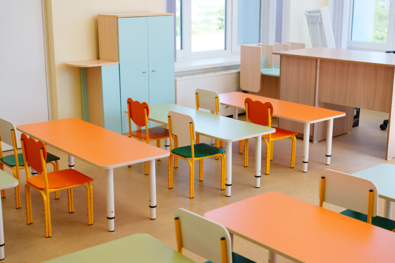 This brightly colored classroom features modern furniture with orange and green tables and chairs. The space is well-lit and designed for interactive learning activities.