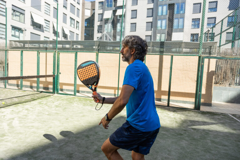 40-year-old paddle player with blue clothes, beard and grey hair, playing a game on a residential paddle court, side view.