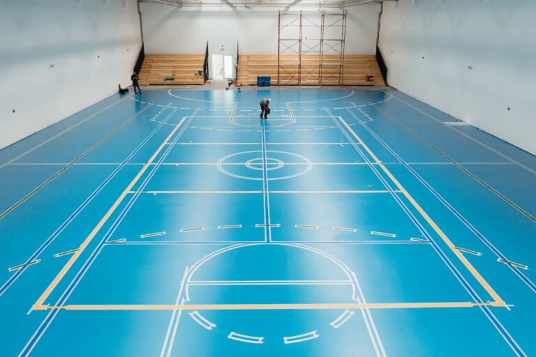 Workers applying epoxy resin on a blue sports court floor in a gymnasium