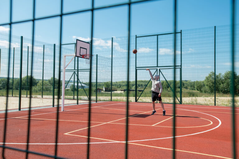 Wide angle portrait of unrecognizable young man throwing ball in hoop while playing basketball alone in outdoor court lit by sunlight, copy space