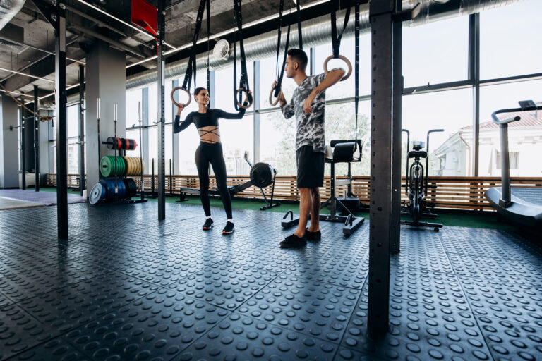 Slim dark-haired girl dressed black sports clothes and athletic man are talking in the spacious gym next to the exercise equipment .