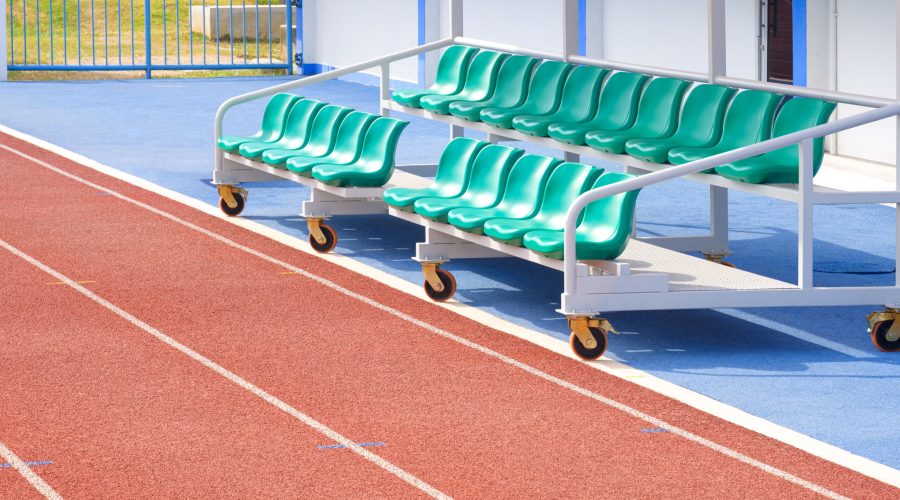 Reserve and coaching staff bench on rubber floor with red synthetic running tracks in outdoor soccer stadium, perspective side view with copy space